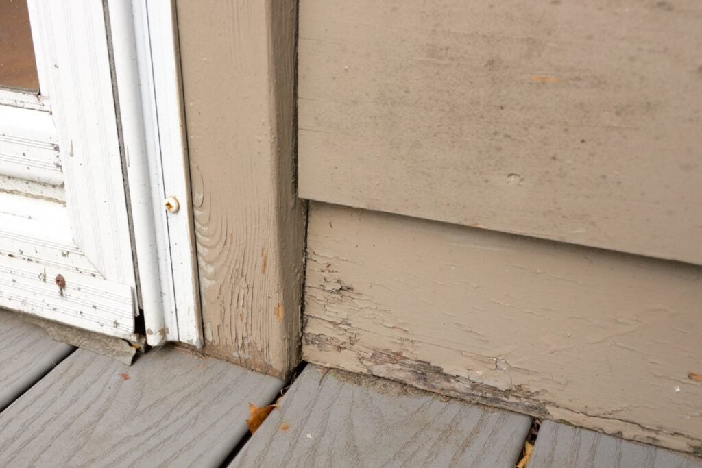 Wood Siding on House shows peeling and cracked painting and early signs of decay and rot where siding meets deck.
