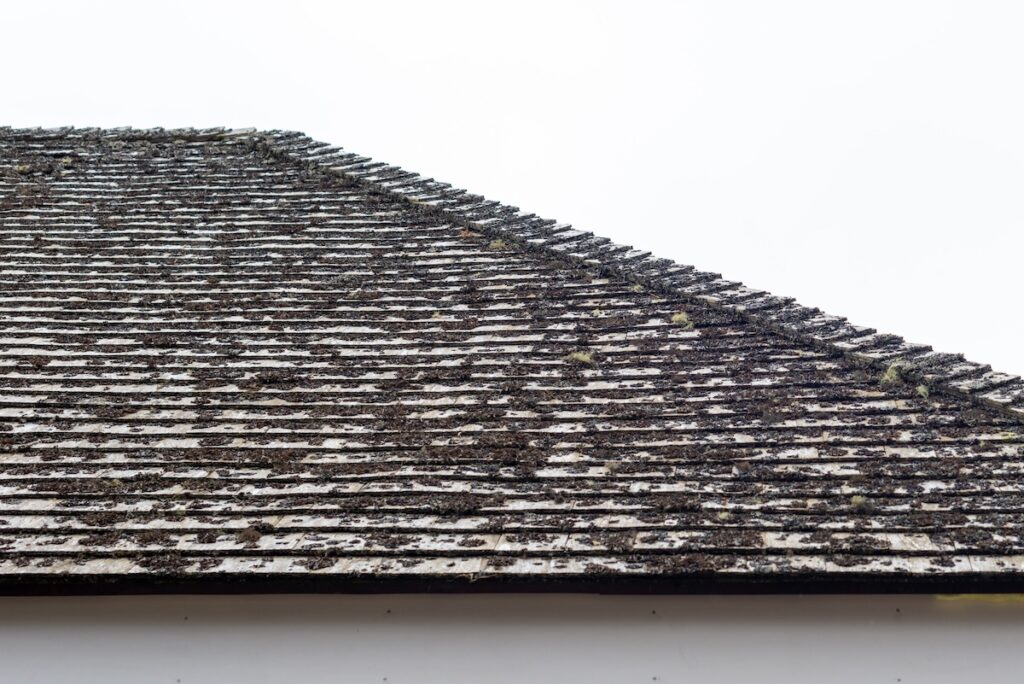A vintage mansard style roof covered in old grey cedar shingles on a house. The material is worn, rough, and has an aged rooftop covering. There's moss among the overlapping layers of roof shingles.